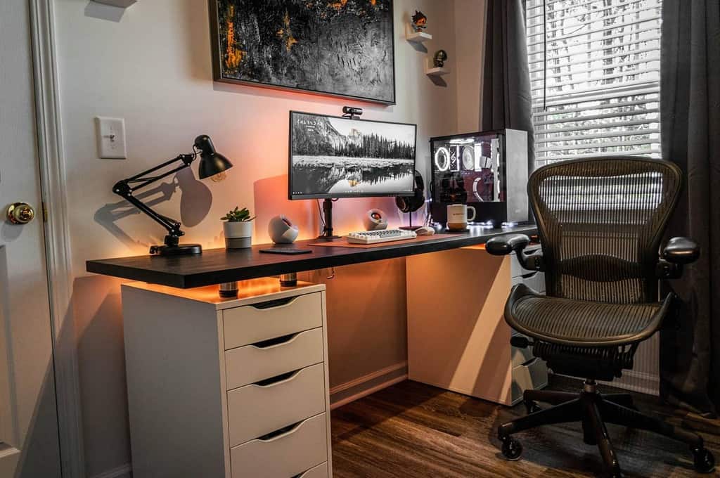 A modern home office with a black desk, ergonomic chair, computer setup, desk lamp, and plant, next to a window with blinds