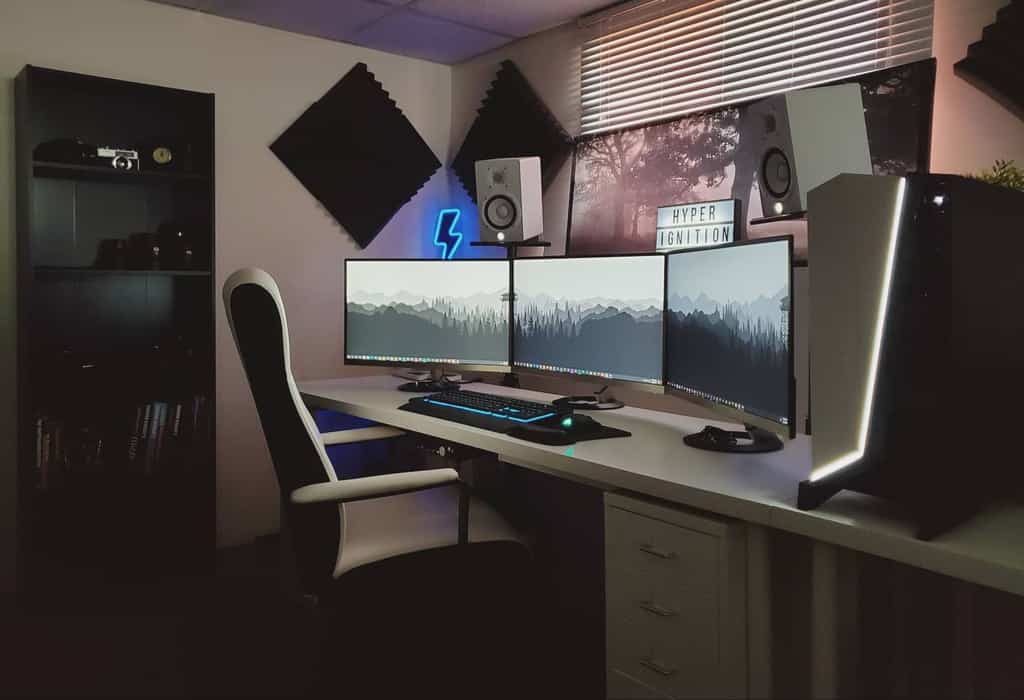 A computer desk setup with three monitors, a keyboard, and speakers, featuring soft lighting and soundproofing panels to create a cozy ambiance