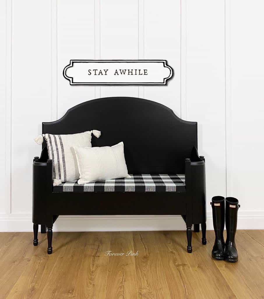 Black chalk-painted farmhouse bench with a buffalo plaid cushion, white accent pillows, and a 'Stay Awhile' sign, styled with rain boots