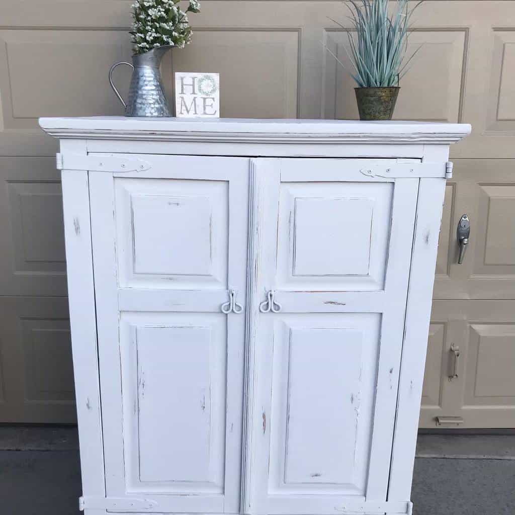 White distressed chalk-painted farmhouse cabinet with rustic metal handles, styled with greenery and a 'Home' sign on top