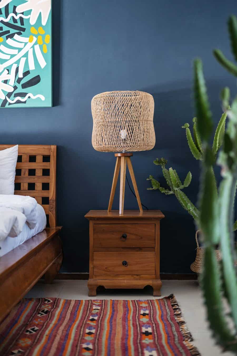 Bedroom corner with a wooden nightstand, wicker lamp, colorful rug, cactus, and abstract wall art against a dark blue wall