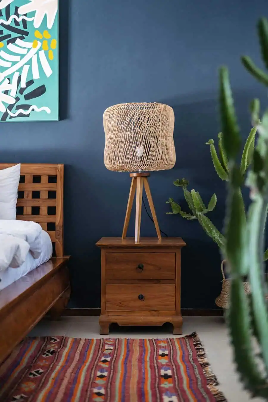 Bedroom corner with a wooden nightstand, wicker lamp, colorful rug, cactus, and abstract wall art against a dark blue wall