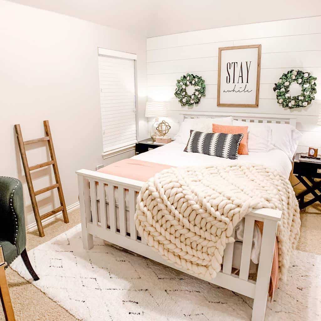 Cozy farmhouse bedroom with a 'Stay Awhile' wood sign, wreath decor, a chunky knit blanket, and a neutral color palette