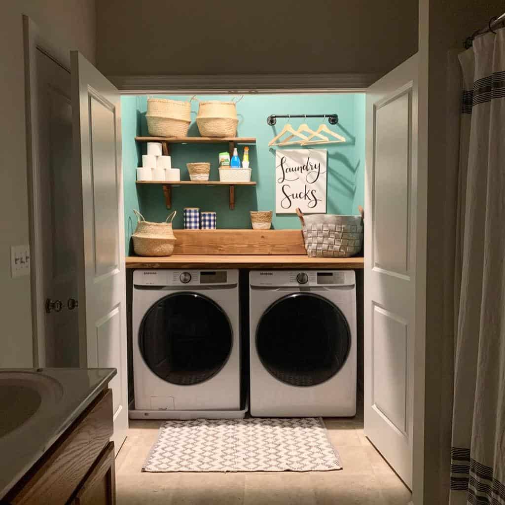 Compact laundry room with washer and dryer, teal wall, shelves, baskets, detergent, and a "Laundry Sucks" sign; cozy setup