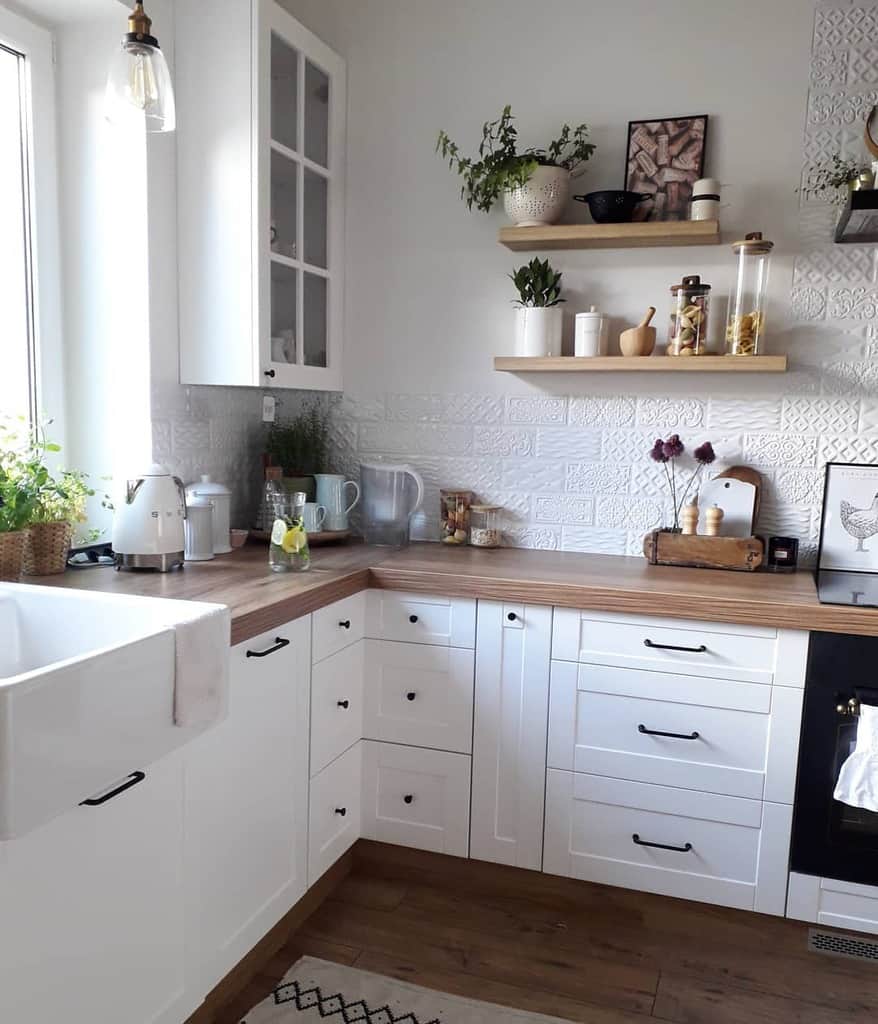 A cozy kitchen with white cabinets, wooden countertops, potted plants, and hanging shelves with jars and decor, light from a large window