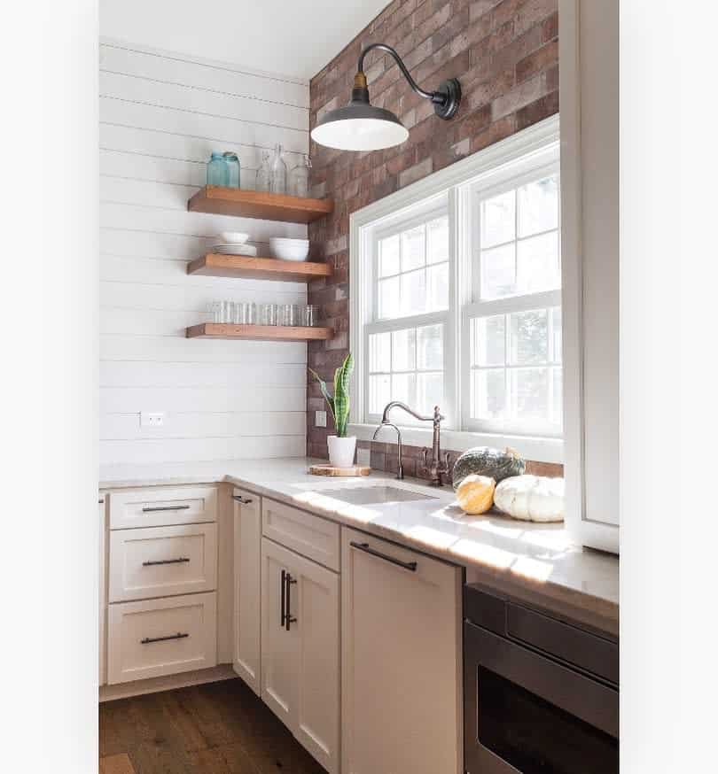 A modern kitchen corner with white cabinets, open wooden shelves, a sink, large window, and a brick accent wall under a wall lamp