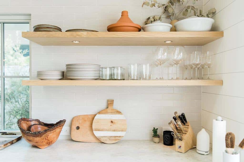 Modern kitchen with wooden shelves holding plates, glasses, and decor; counter has cutting boards, a knife block, and a small plant