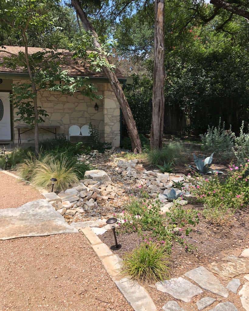 Rustic front yard xeriscape with native plants, gravel pathways, a dry creek bed, and stone accents surrounding a cozy stone house