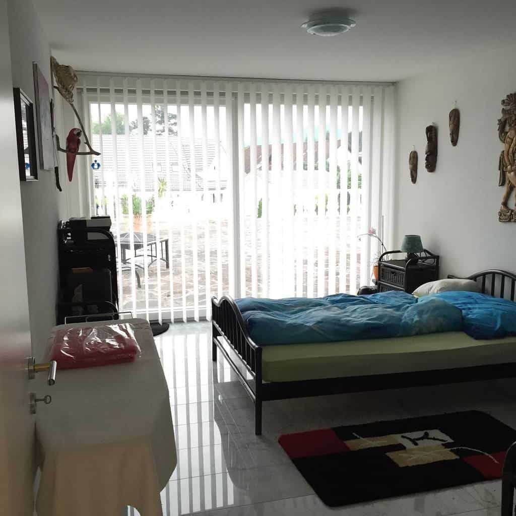 Minimalist bedroom with a single bed, patio view through vertical blinds, decorated walls, and white tiled floor