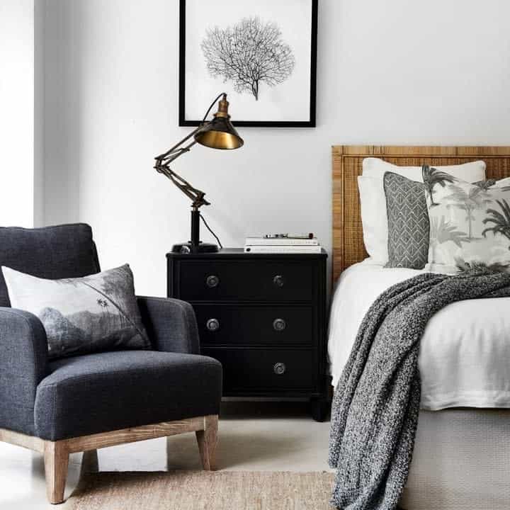 Cozy bedroom with a grey armchair, wooden bed, patterned pillows, textured blanket, black nightstand, and a framed tree artwork