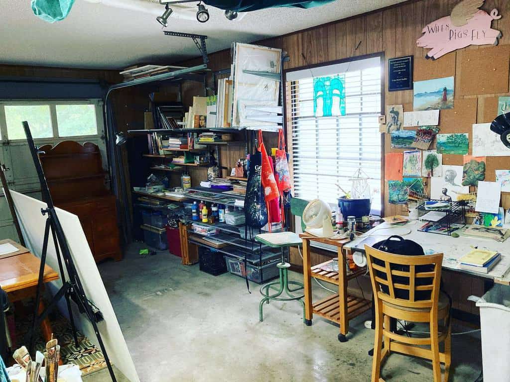 Cozy garage art studio with wooden walls, open shelving, a drafting table, and creative works pinned on a corkboard