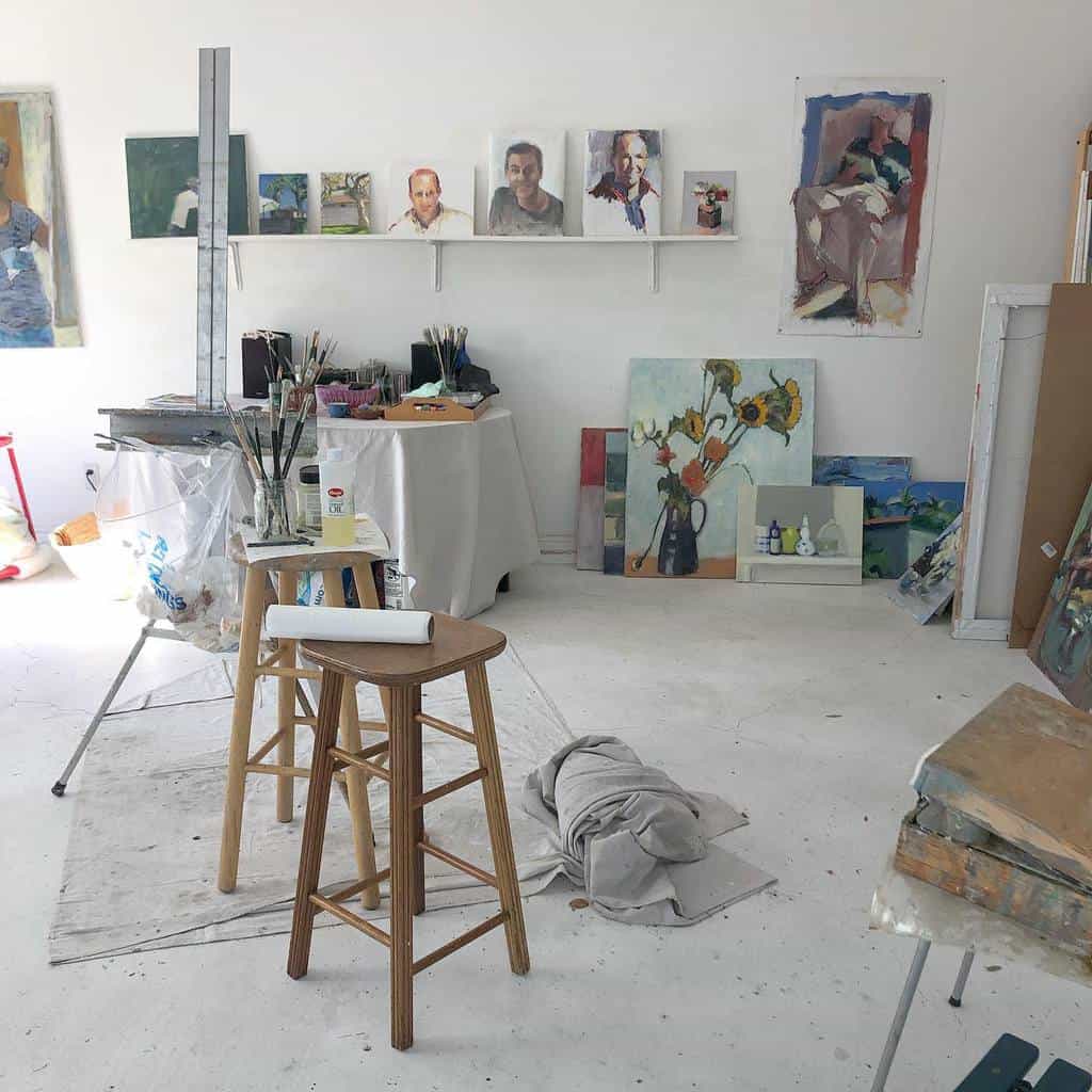 Art studio with paintings on walls and floor, stools, easel, brushes, and a draped cloth on the ground in a bright, airy atmosphere