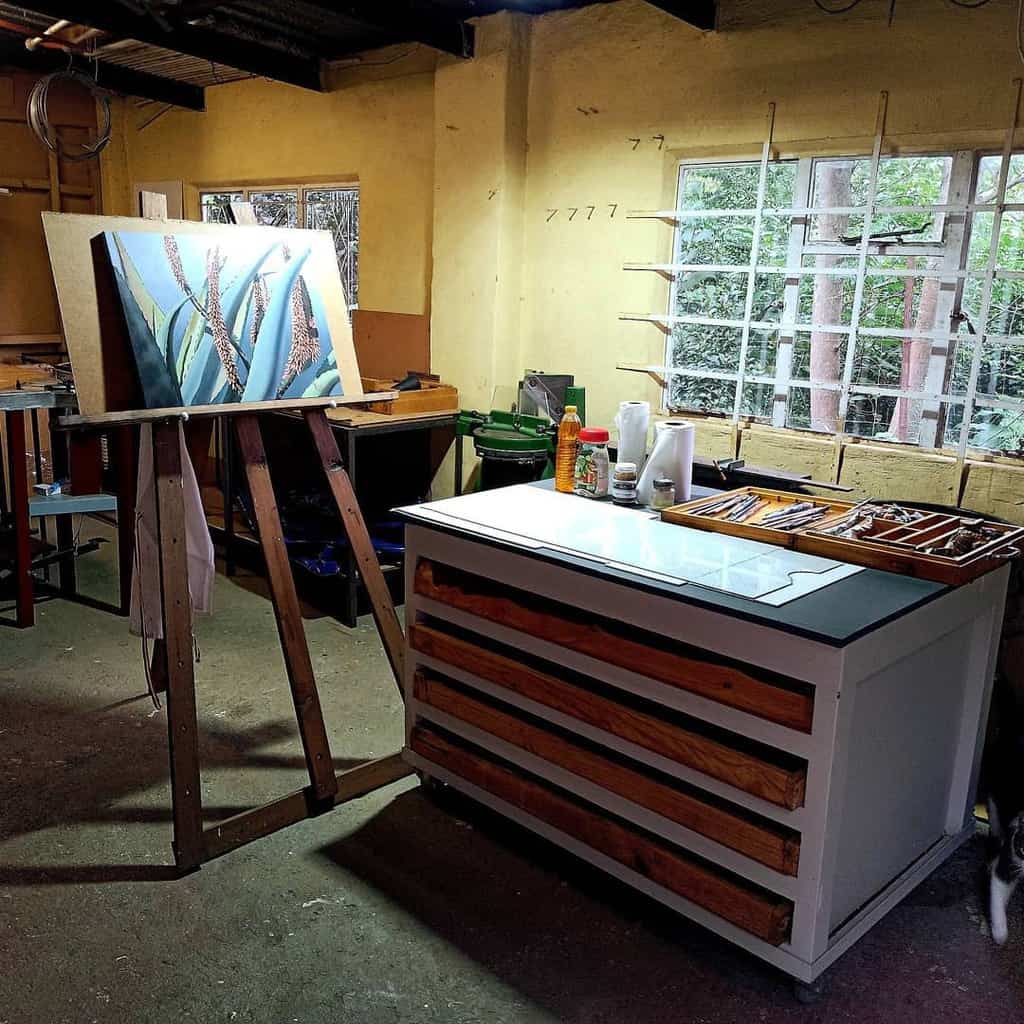 An artist's studio with a canvas of plants on an easel, a cabinet with drawers, and art supplies near a window