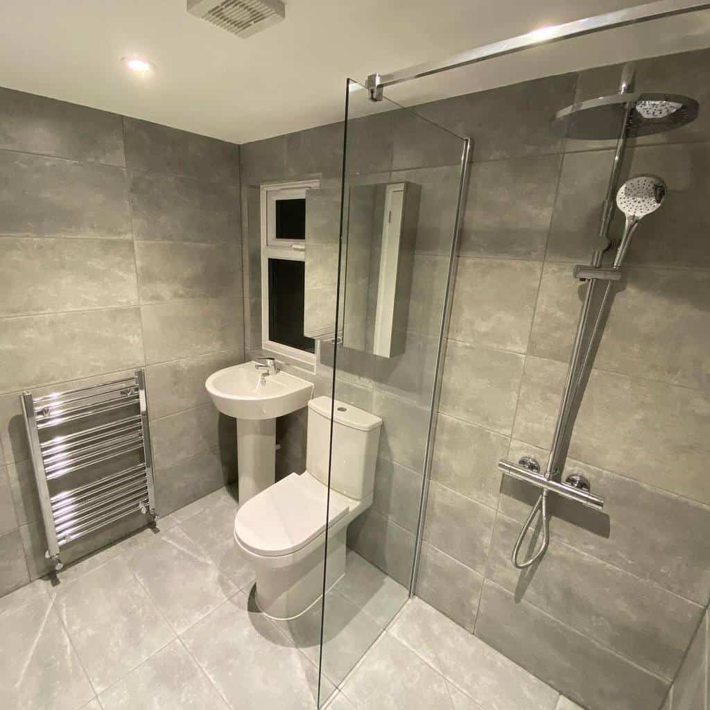 Modern wet room with gray tiles, a glass shower, white pedestal sink, toilet, and wall-mounted towel rack; ceiling lights provide illumination