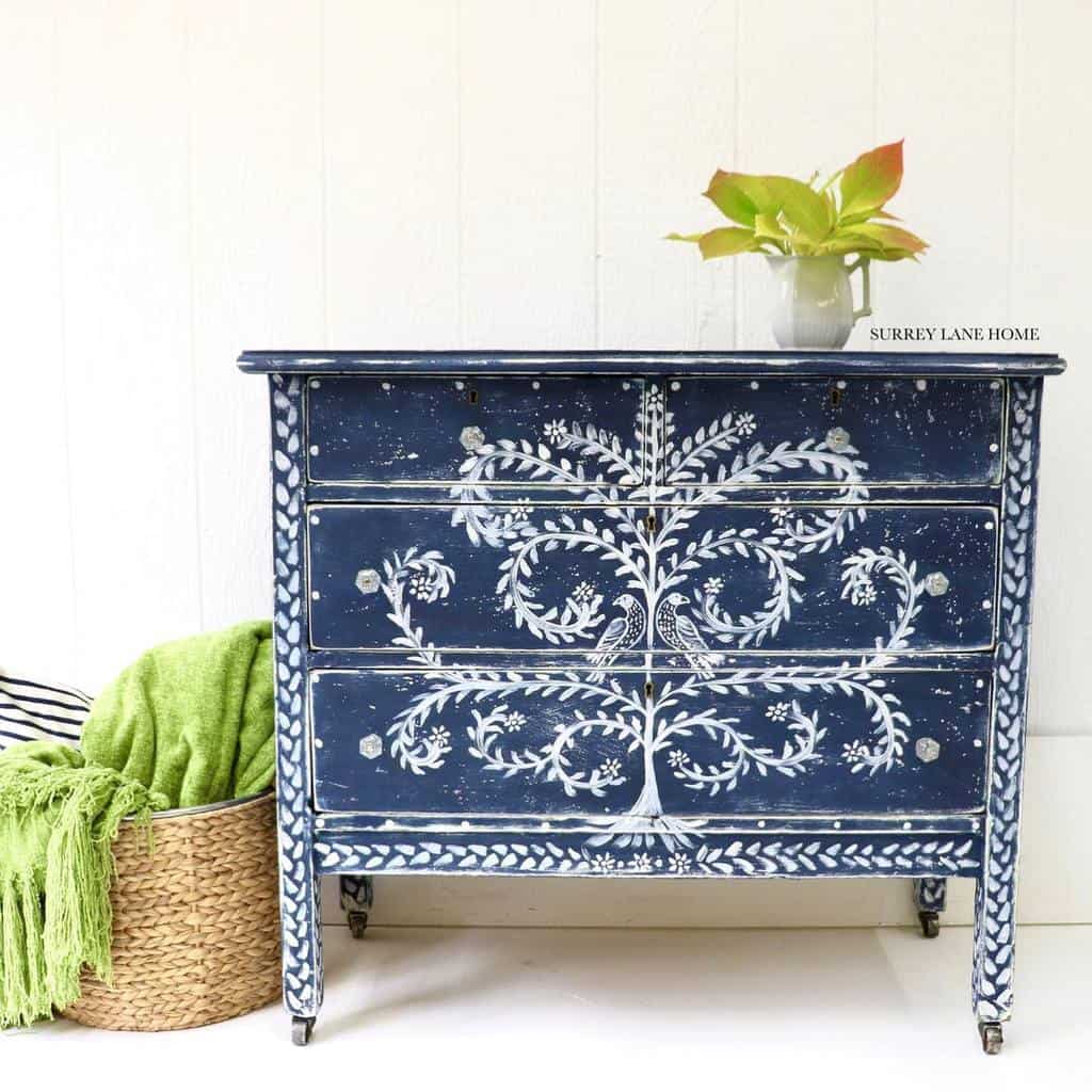 Vintage navy blue chalk-painted dresser with intricate hand-painted white floral designs, styled with a plant and woven basket