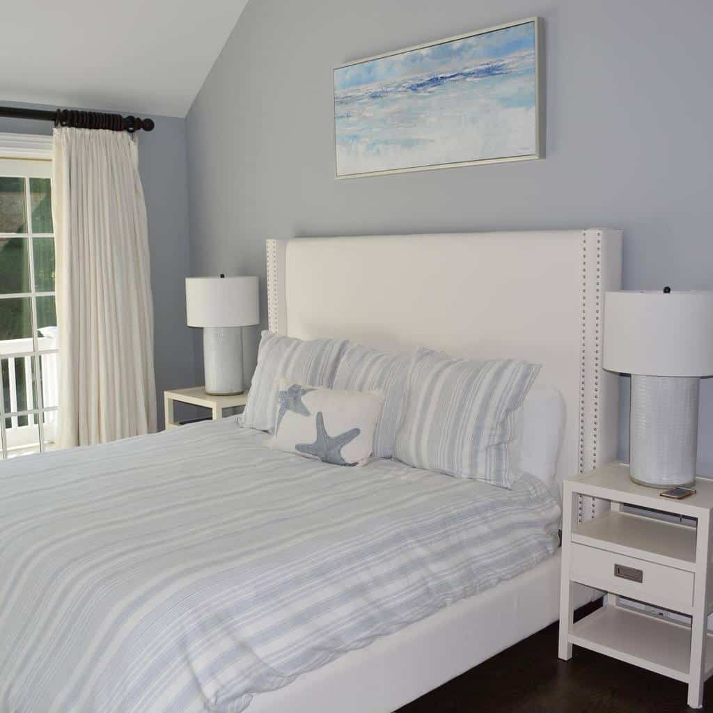 A bedroom with a white bed, striped bedding, starfish cushions, two white nightstands with lamps, and a beach painting on the wall