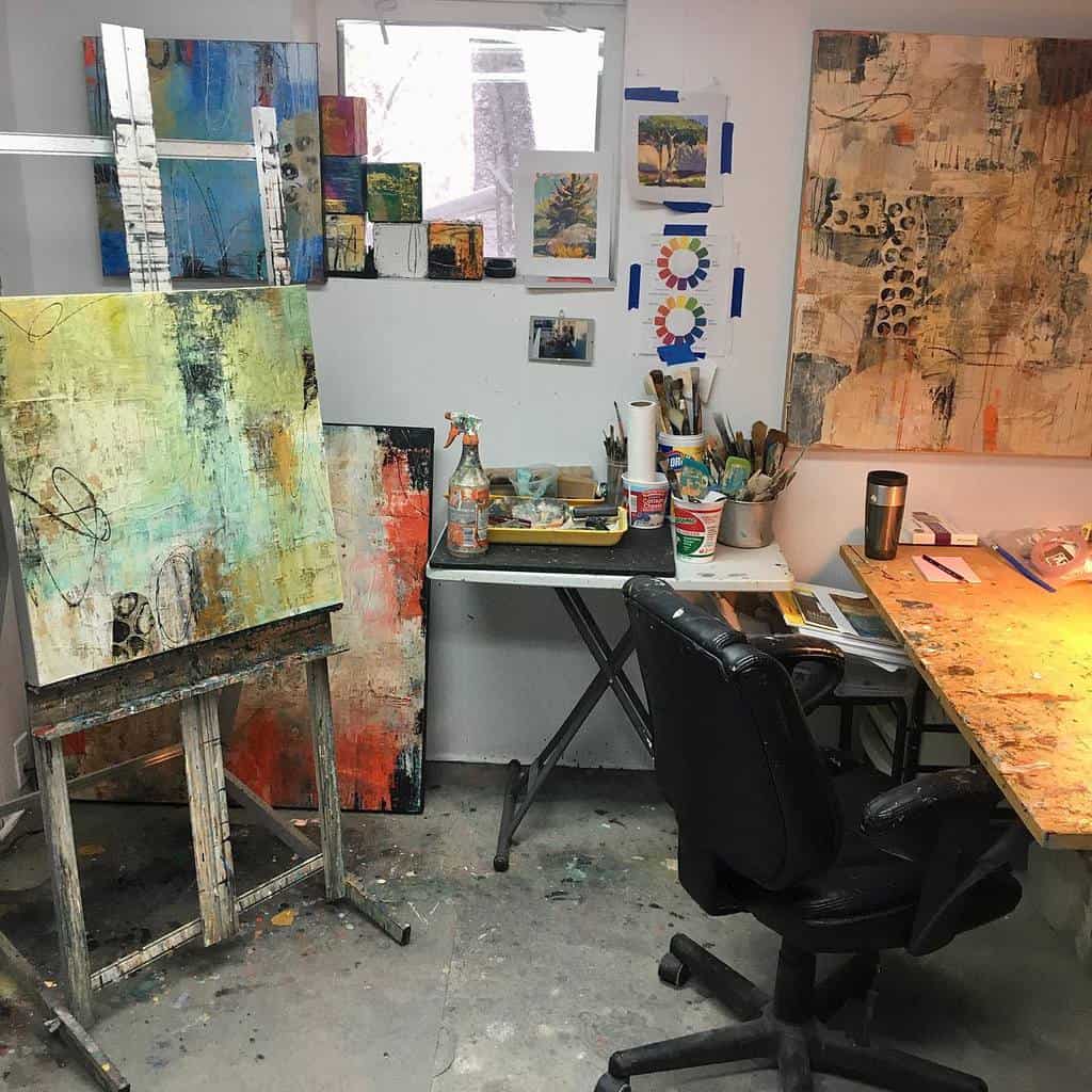 Art studio with easels displaying abstract paintings, a cluttered desk with art supplies, and a chair in front of a work table