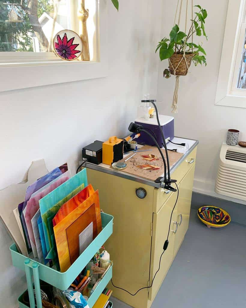 Cozy art corner with glass-cutting tools, colorful paper stack, hanging plant, and window; a warm and inviting space for creativity