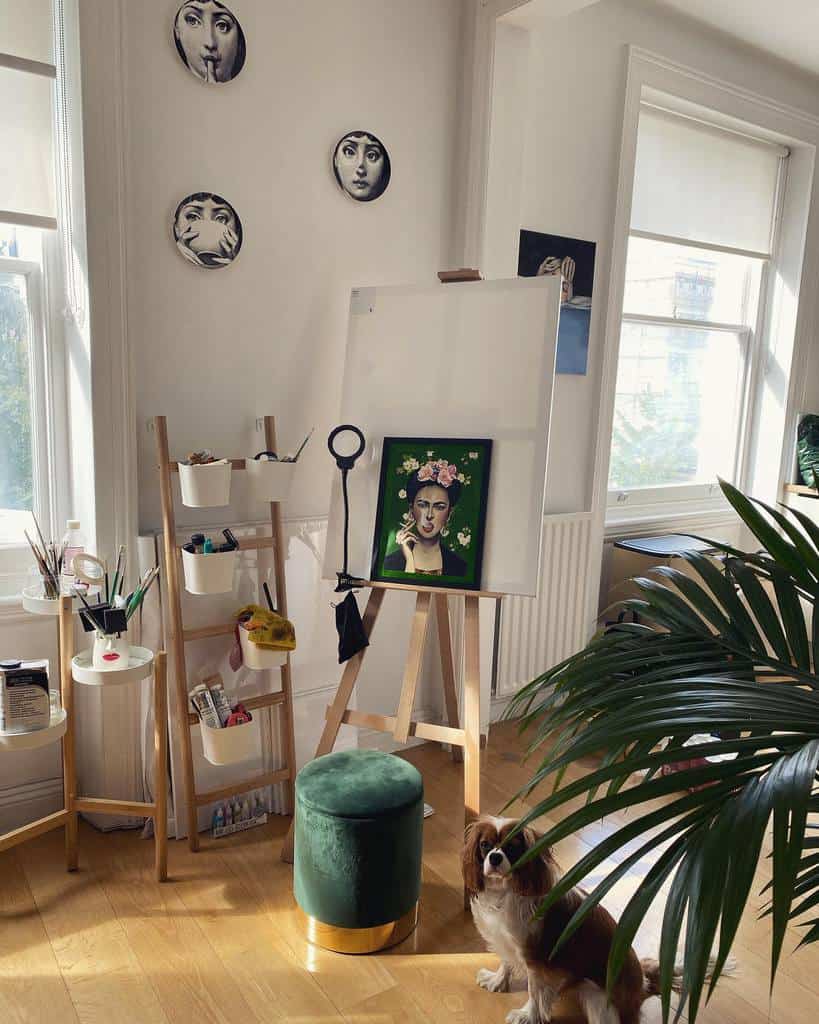 Bright home art studio with a wooden easel, creative wall decor, organized art supplies, a green velvet stool, and a small dog