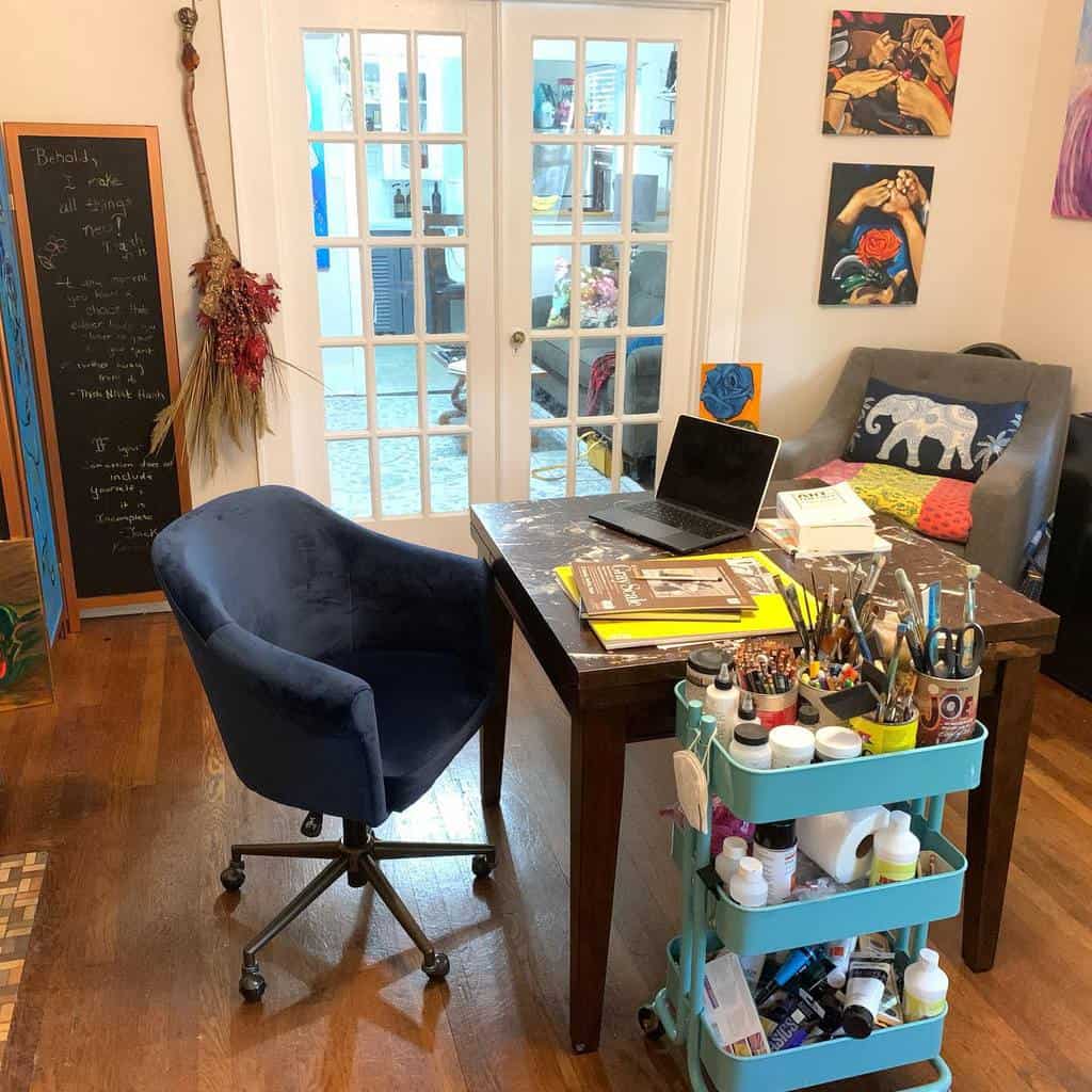 Cozy art studio with a wooden desk, blue chair, art supplies on a cart, and a couch with a decorative elephant pillow in the background