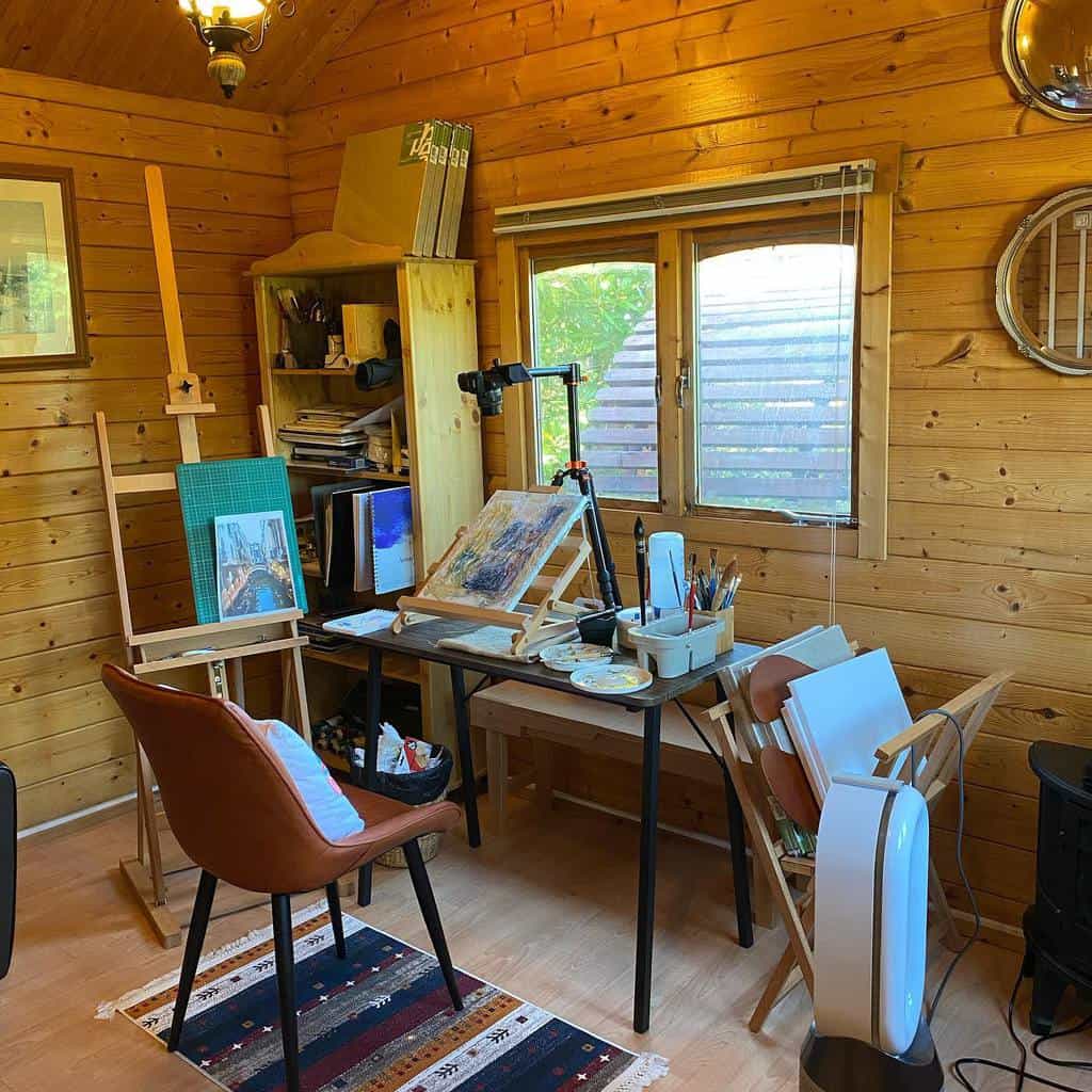 Cozy art studio with wooden walls, easels, painting supplies, a chair, and a rug; a window lets in natural light