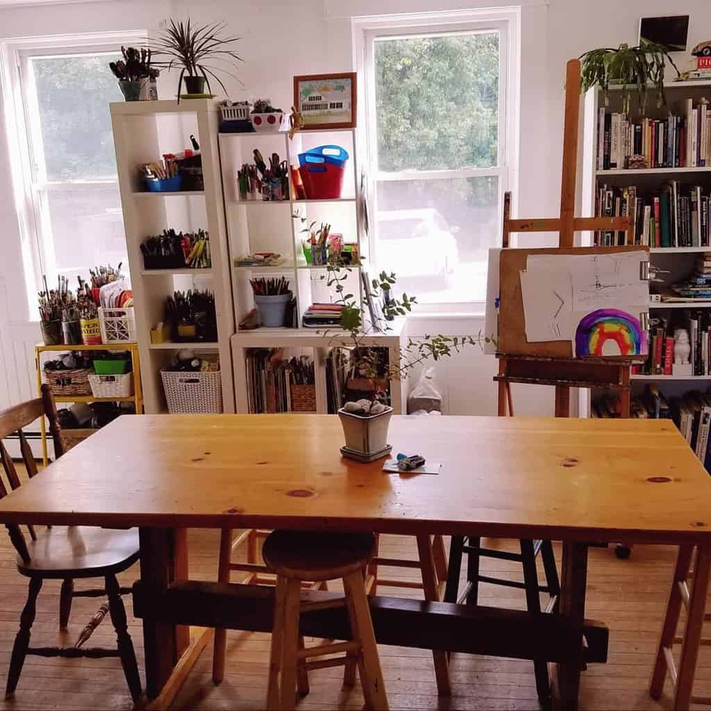 Art studio with a wooden table surrounded by chairs and stools, shelves with art supplies, and a painting on an easel near two windows