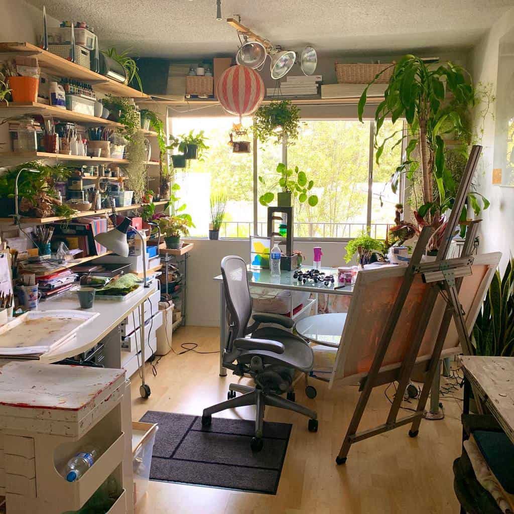 A sunny art studio filled with plants, a drafting table, shelves of supplies, and a chair creates a vibrant and creative atmosphere