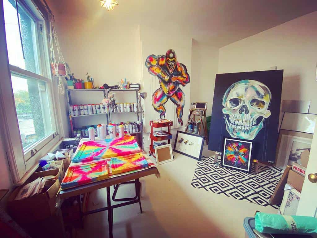 Art studio with colorful murals of a skull and a jumping figure, spray paint cans on shelves, and tie-dye fabric on a table