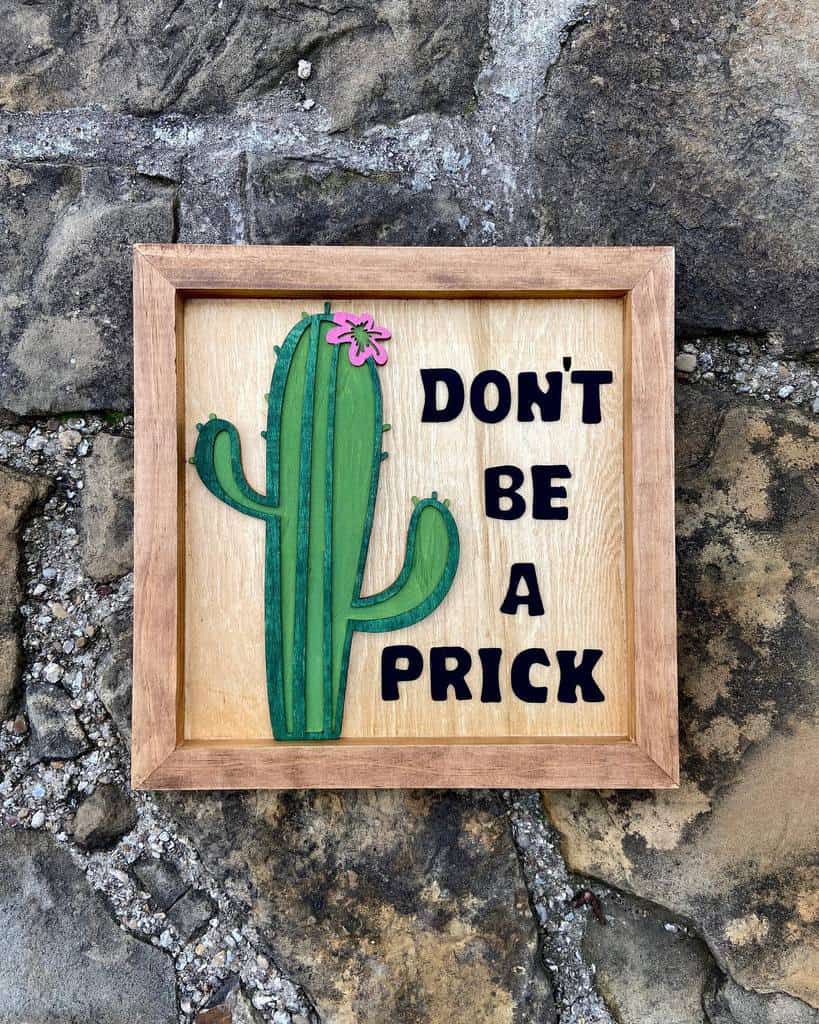 Wooden sign with a cactus and a pink flower, reads "DON'T BE A PRICK," placed on a stone surface