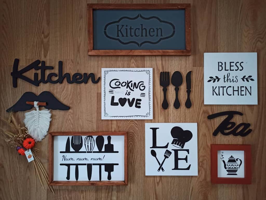 Wooden kitchen wall decorated with black and white farmhouse-style signs, including 'Bless This Kitchen' and 'Cooking is Love,' with utensil designs