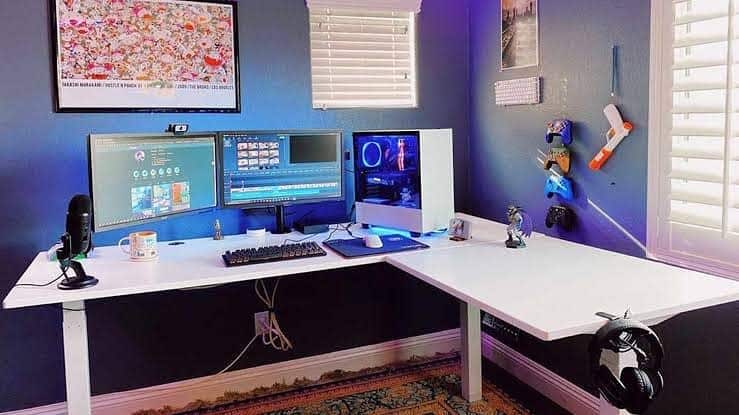 A corner desk setup with dual monitors, a white PC, microphone, keyboard, mouse, and wall-mounted gadgets in a blue room with posters