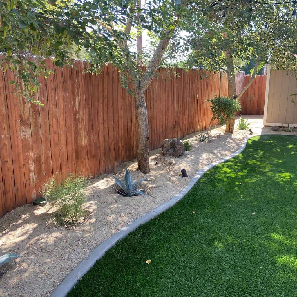 Backyard xeriscape with drought-tolerant plants, trees, a wooden fence, artificial turf, and a gravel border for a clean, modern look