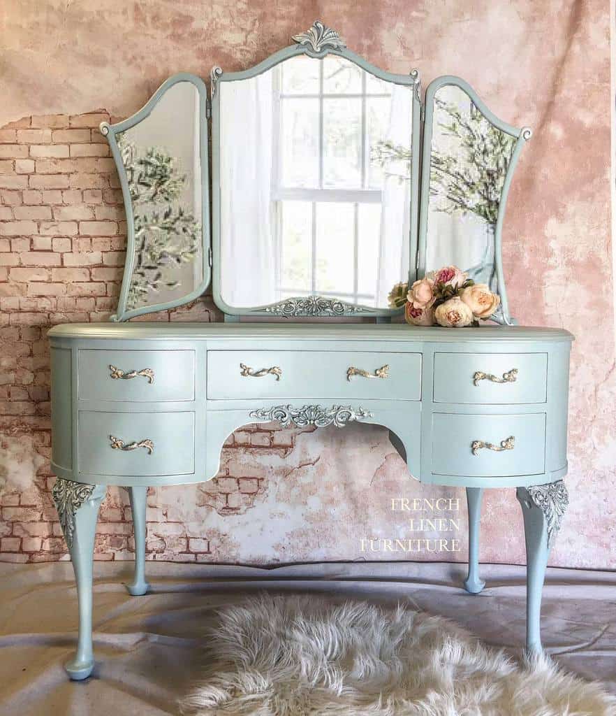 Elegant vintage vanity in soft blue chalk paint with ornate gold handles and a tri-fold mirror, styled with roses and a faux fur rug