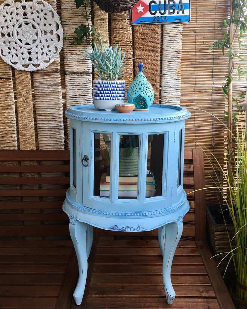 Light blue chalk-painted vintage display cabinet with glass panels, styled with boho decor, potted plants, and books on a wooden patio