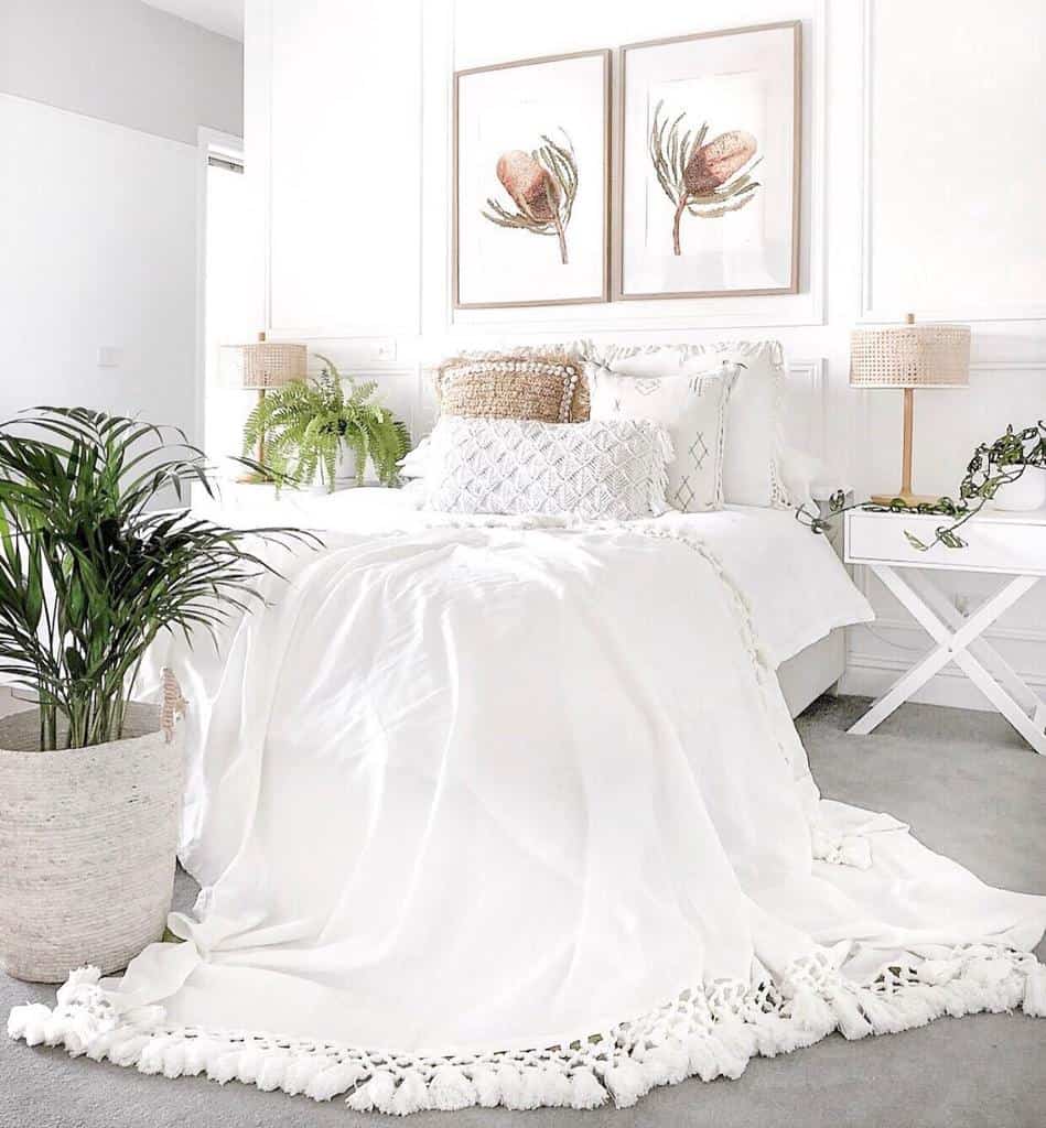 Bright bedroom with a white bed, tassel blanket, potted plants, and botanical wall art, featuring neutral decor and soft lighting