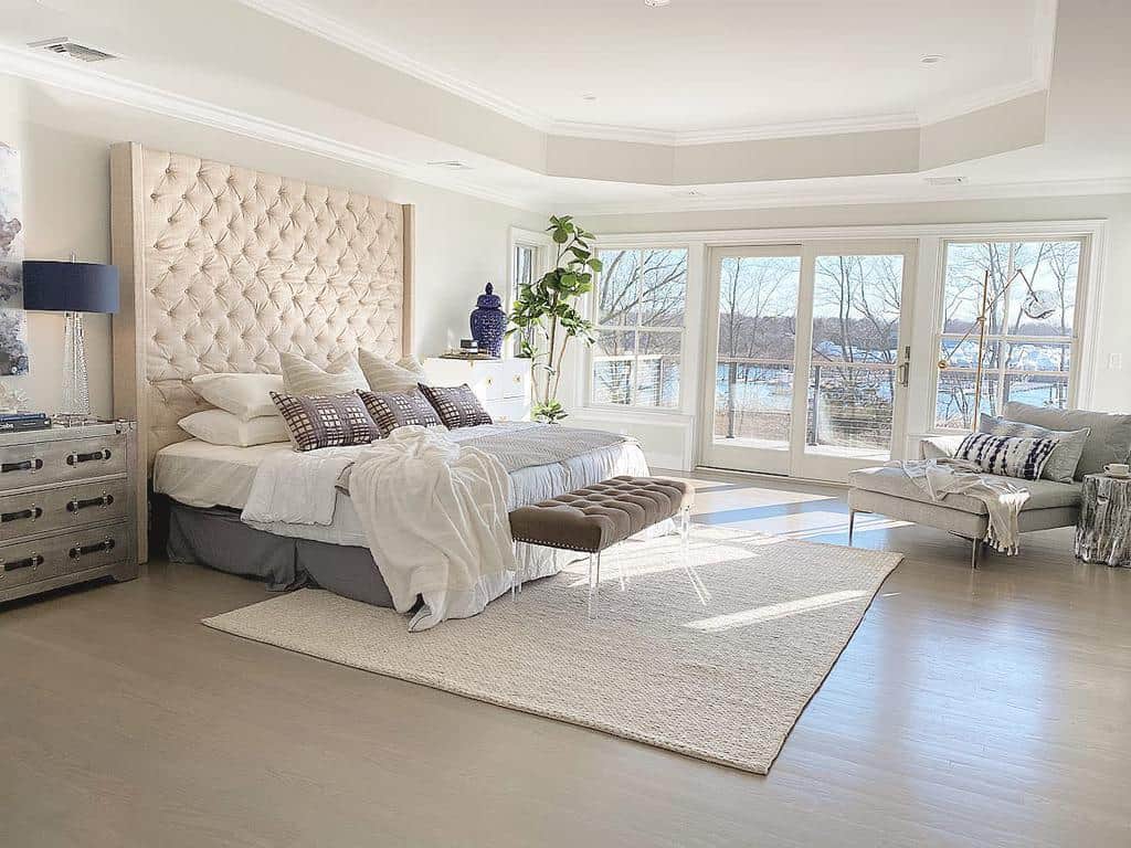 Luxurious bedroom with a large tufted headboard, gray bedding, and a view of a lake through large windows, bright and spacious decor