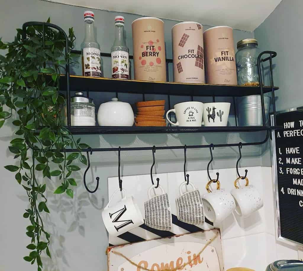 Kitchen shelf with drink syrups, mugs, plants, and tins labeled "Fit Chocotella" and "Fit Vanilla," with mugs hanging underneath on hooks