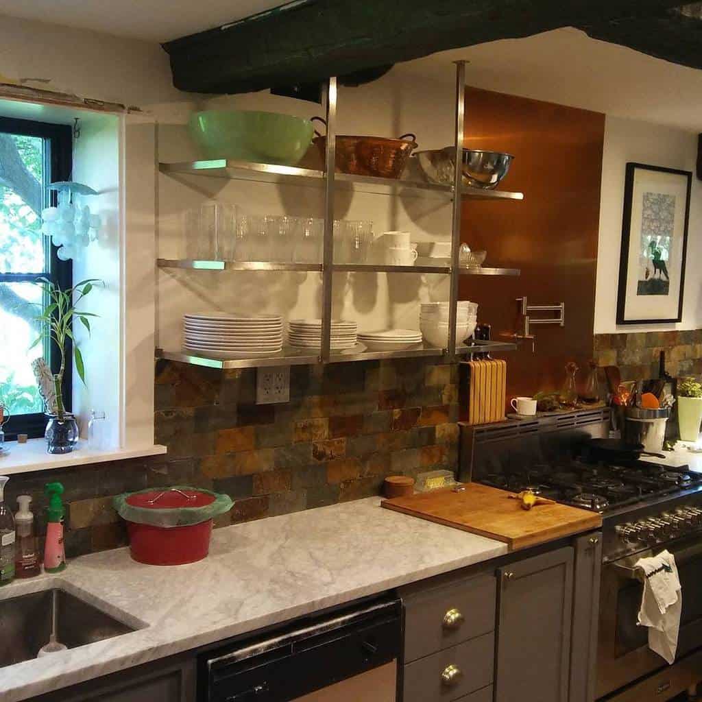 A kitchen with marble counters, dark cabinets, open metal shelves with dishes, and a stove featuring a brick backsplash accentuating the space