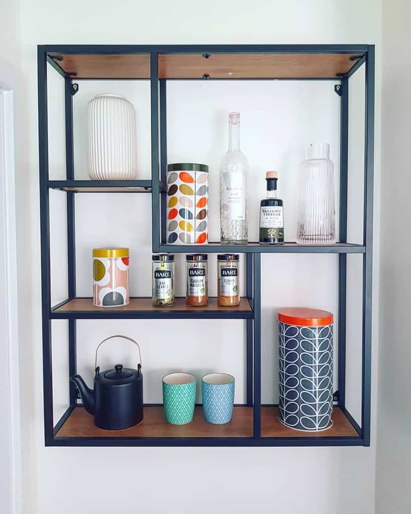 Wall-mounted shelf with a black frame holding decorative canisters, three bottled drinks, a black teapot, and two blue mugs