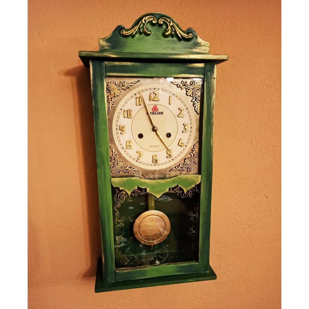 Vintage green chalk-painted wall clock with gold metallic accents, ornate detailing, and a glass panel showcasing the swinging pendulum