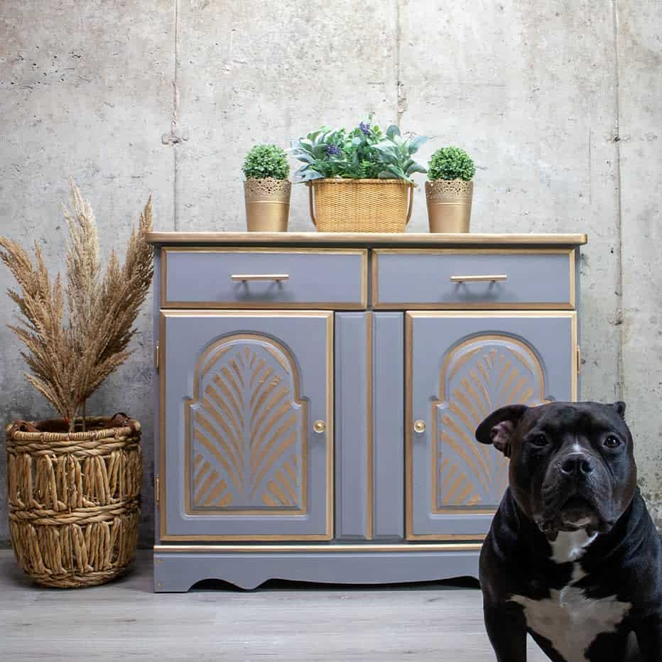 Gray chalk-painted sideboard with gold metallic detailing, arched panel designs, and potted greenery, styled with a woven basket and a dog