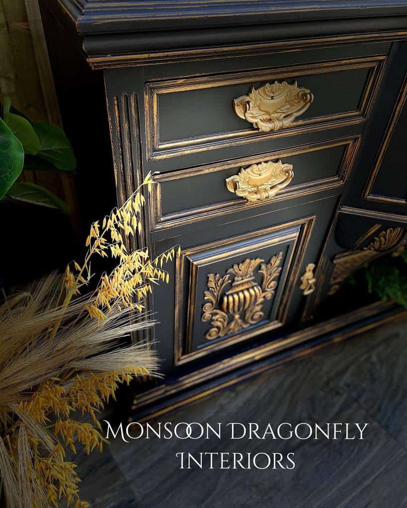 Black chalk-painted vintage cabinet with ornate gold metallic detailing and intricate hardware, styled with dried wheat and greenery