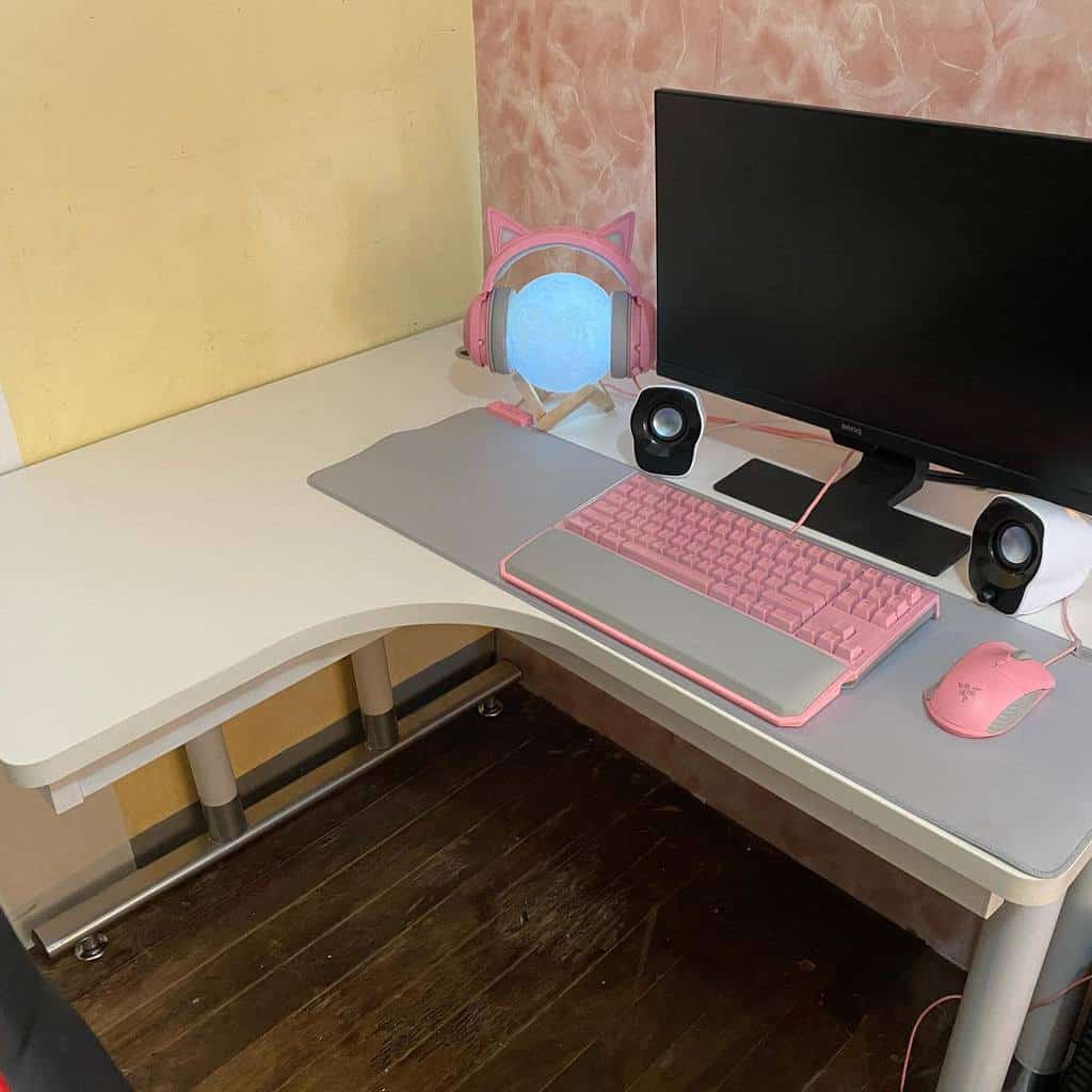 L-shaped desk with a computer, pink and gray keyboard, pink mouse, pink cat ear headphones, and speakers on a wooden floor