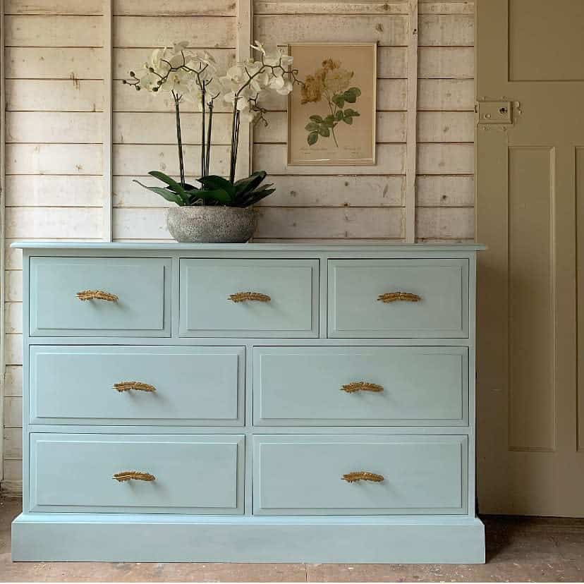 Light blue chalk-painted dresser with gold textured handles, styled with a potted orchid, botanical artwork, and a rustic wooden backdrop