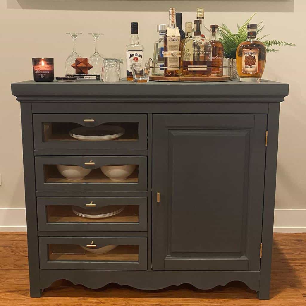 Matte charcoal chalk-painted sideboard with glass-front drawers and gold hardware, styled as a bar with liquor bottles and glassware