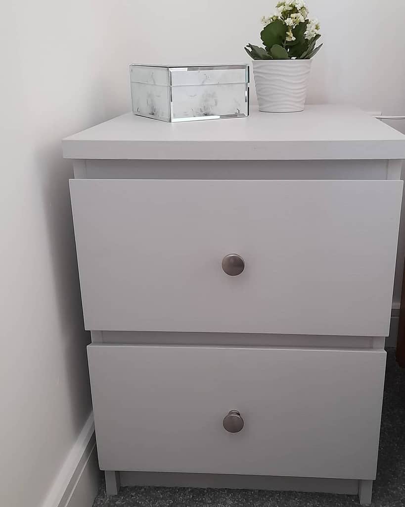 Minimalist light gray chalk-painted nightstand with round metal knobs, styled with a mirrored jewelry box and a potted plant