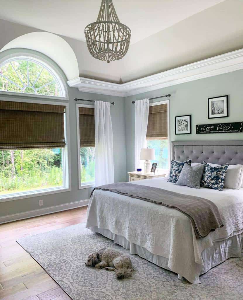 Bright bedroom with large arched window, white curtains, and a bed with gray accents. A dog lies on a patterned rug