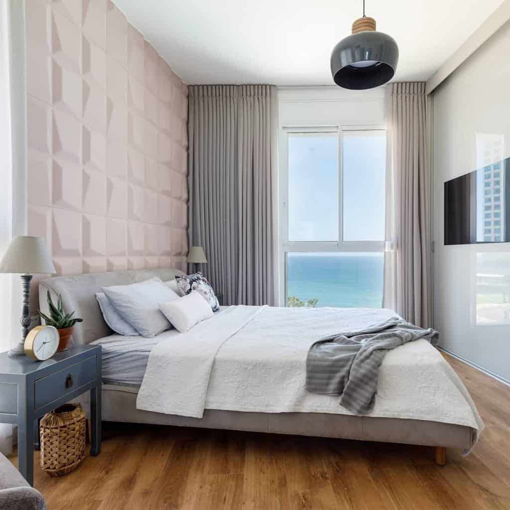 Modern bedroom with a large bed, pale pink accent wall, ocean view window, gray curtains, wooden floor, and blue nightstand