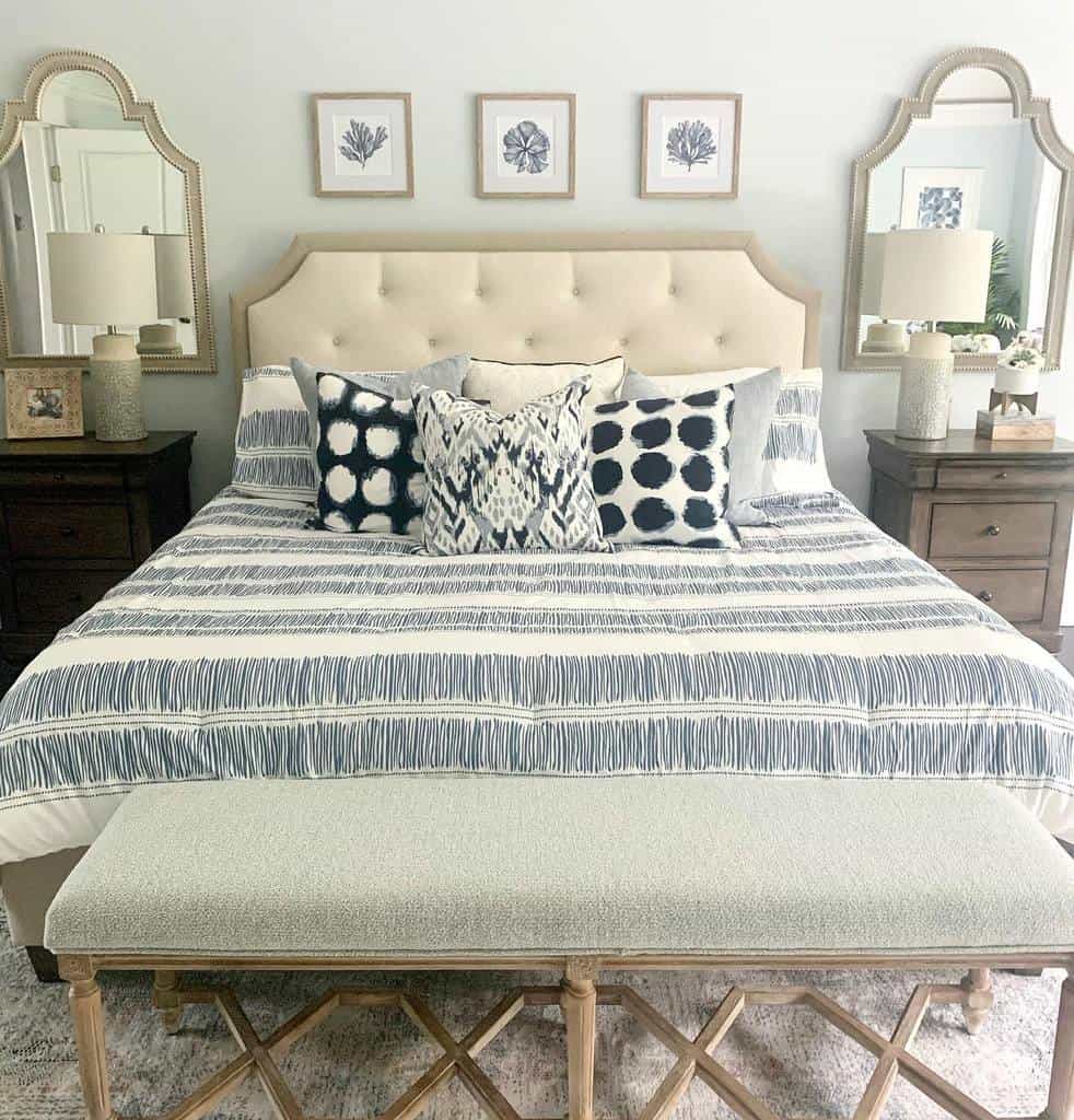 Bedroom with a beige upholstered bed, patterned pillows, and a bench; nightstands with lamps and four framed pictures above the headboard