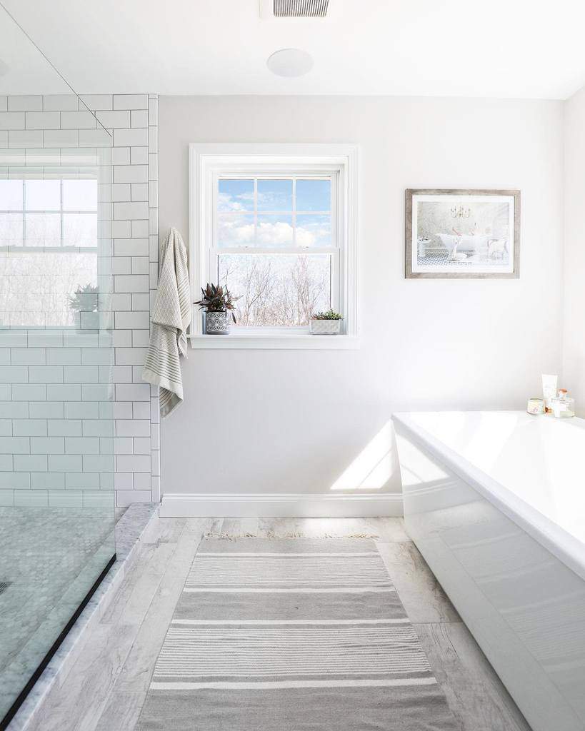 Bright wet room with a window, white tub, glass shower, striped rug, two plants, and a towel hanging on the tile wall